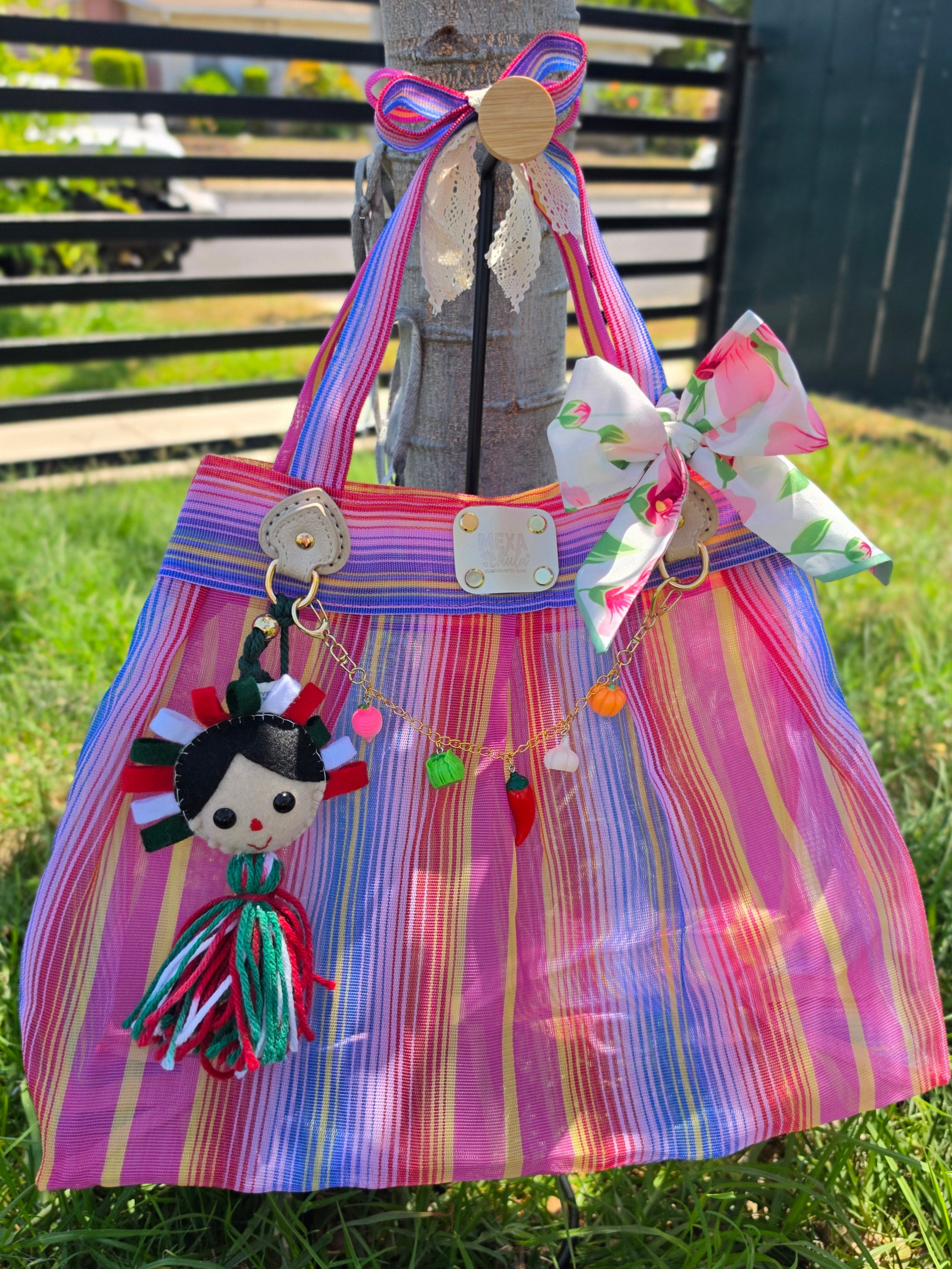 Bolsa de mercado mexicana de rayas rosa, azul y amarillo con charm de muñequita y moño floral rosa colgada de un árbol