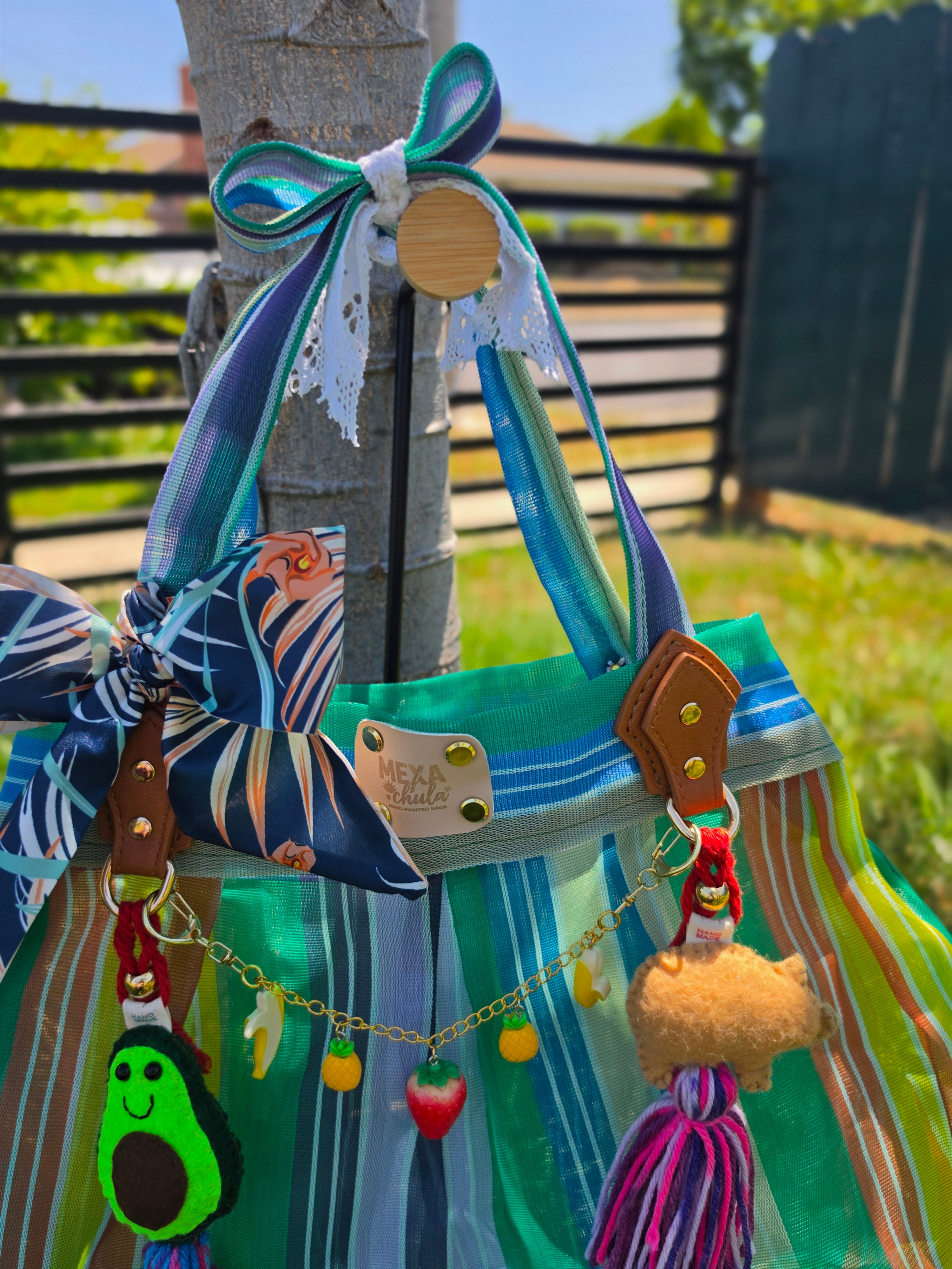Bolsa de mercado mexicana rayas verde, azul y coral con charms de aguacate y cerdito de fieltro colgada de un árbol